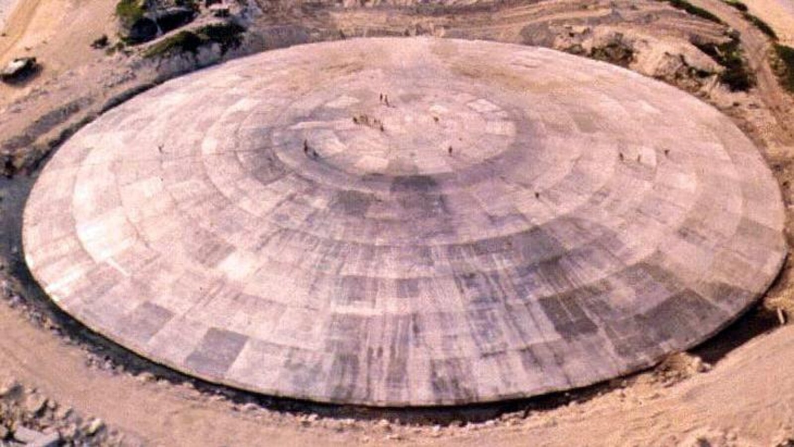 Un vaste dôme circulaire de béton trône sur une petite île verte encerclée d'eau bleue.