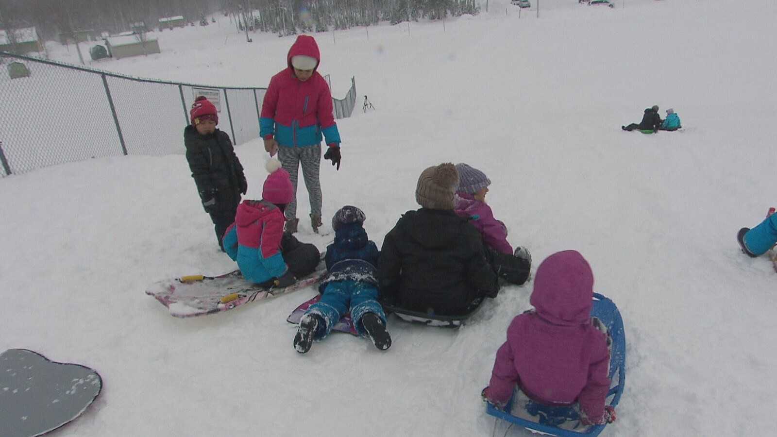Des enfants, accompagnés d'adultes, descendent une pente de neige sur des planches.