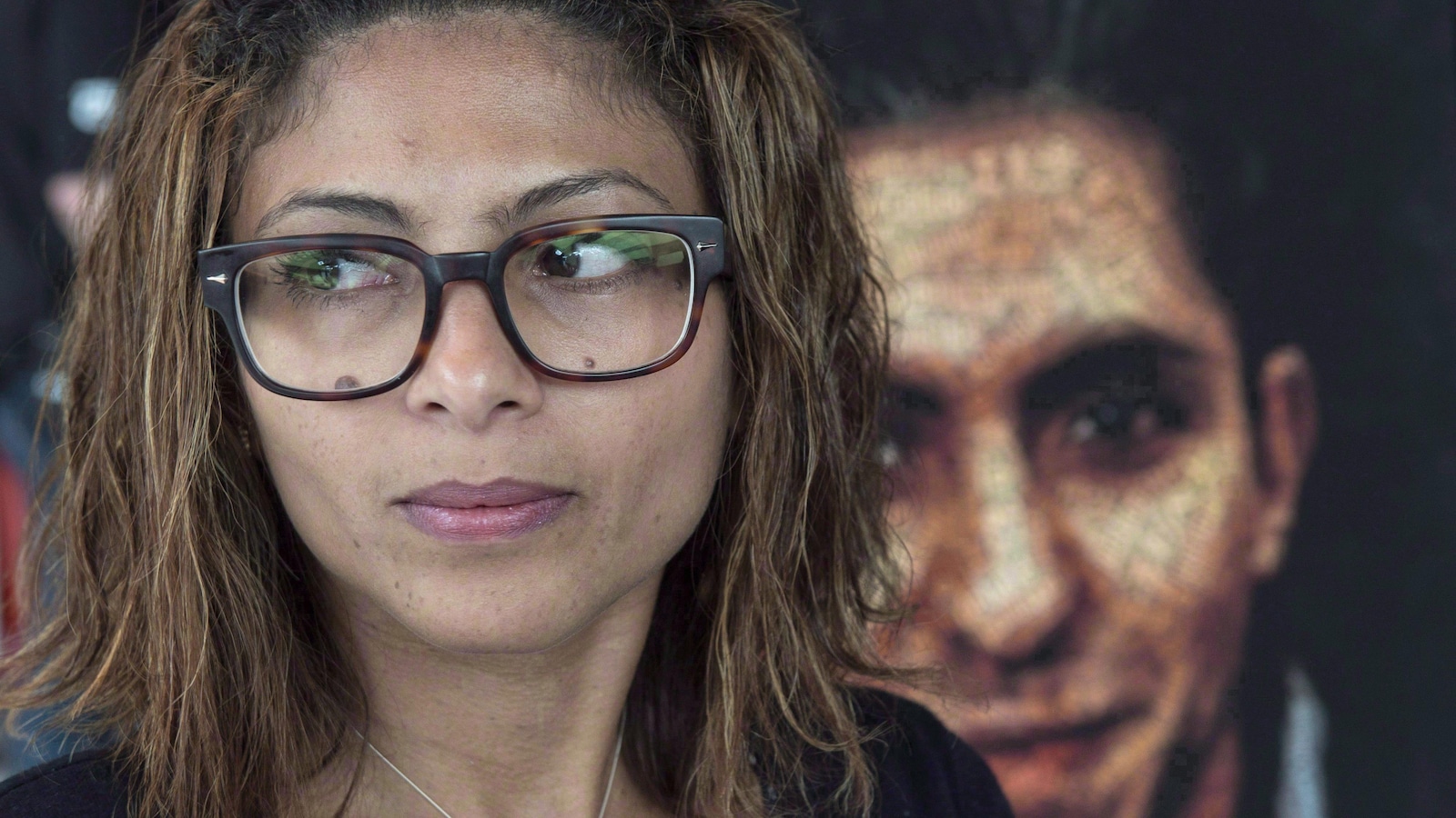Une femme portant des lunettes carrées regarde de biais devant le portrait de son mari.