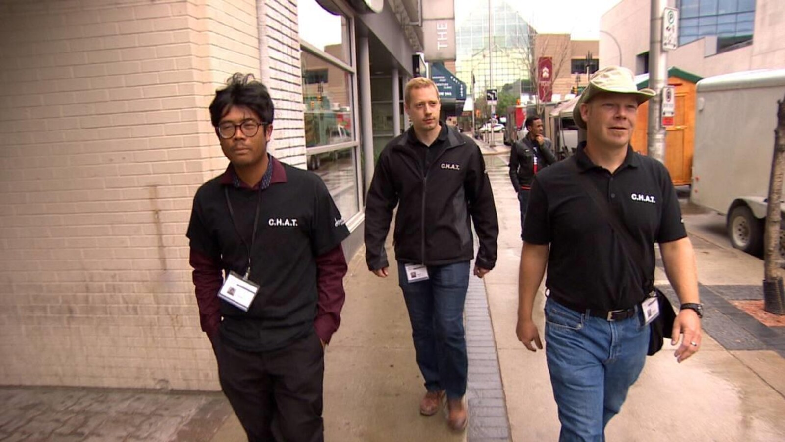 Trois hommes portant des chandails et des manteaux noirs avec le sigle C.H.A.T. marchent sur le trottoir par une journée pluvieuse.