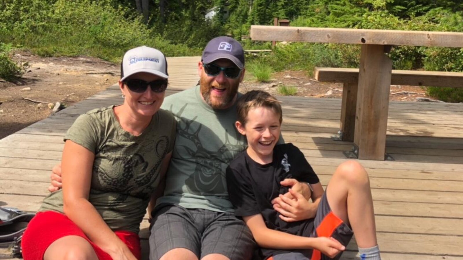 Une famille souriante assise sur une plateforme en bois dans un sentier pédestre dans la forêt.