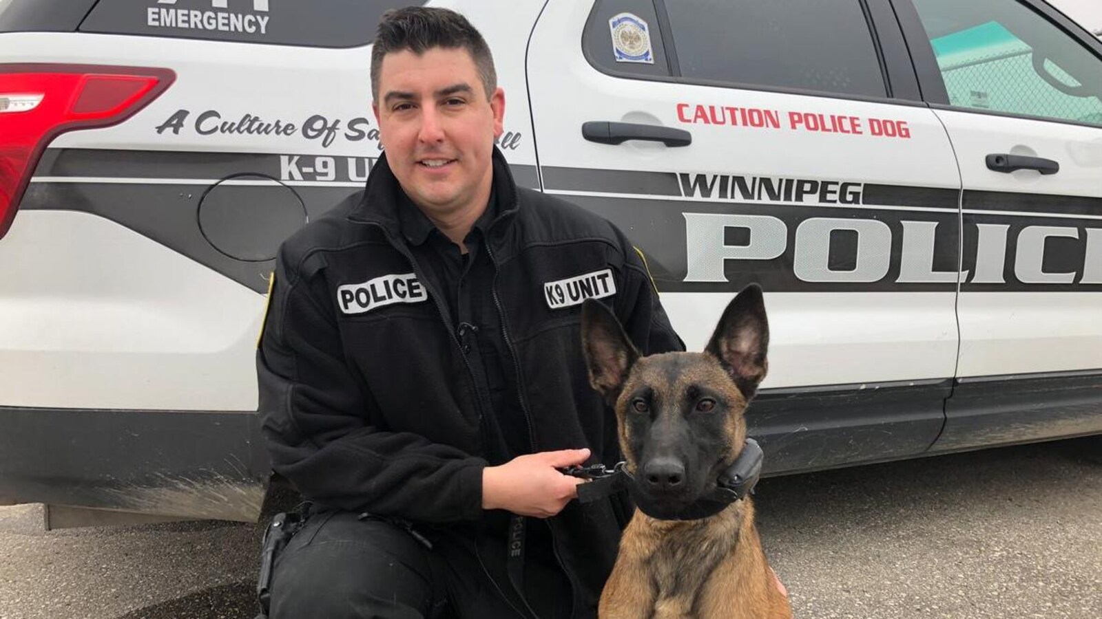 Un policier tient un chien en laisse devant une voiture de police.