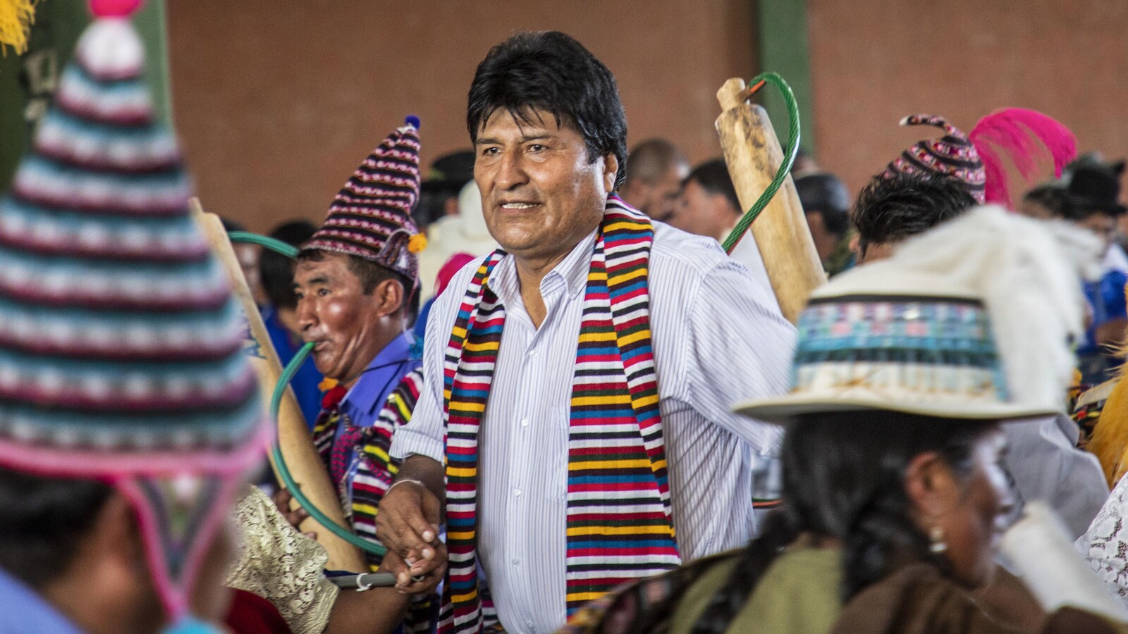 Un homme portant une chemise aux manches retroussées et un foulard multicolore danse avec des Boliviens en tenues traditionnelles.