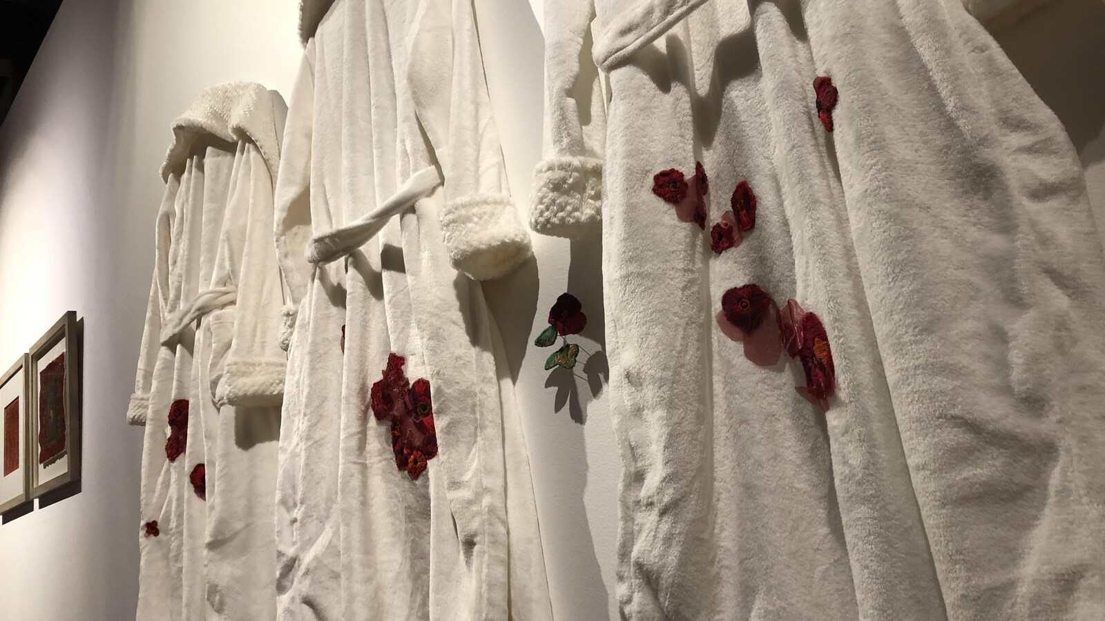 Trois robes de chambre sont accrochées sur un mur à coté de tableaux. De fausses fleurs sont collées sur les vêtements.