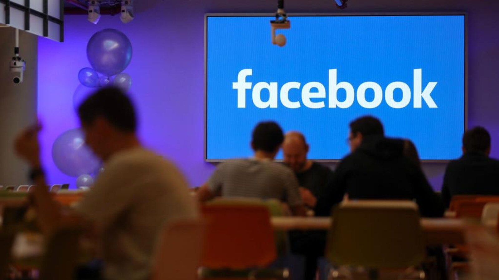 Des hommes déjeunent à table dans une grande salle, avec en fond un écran bleu affichant le logo de Facebook.
