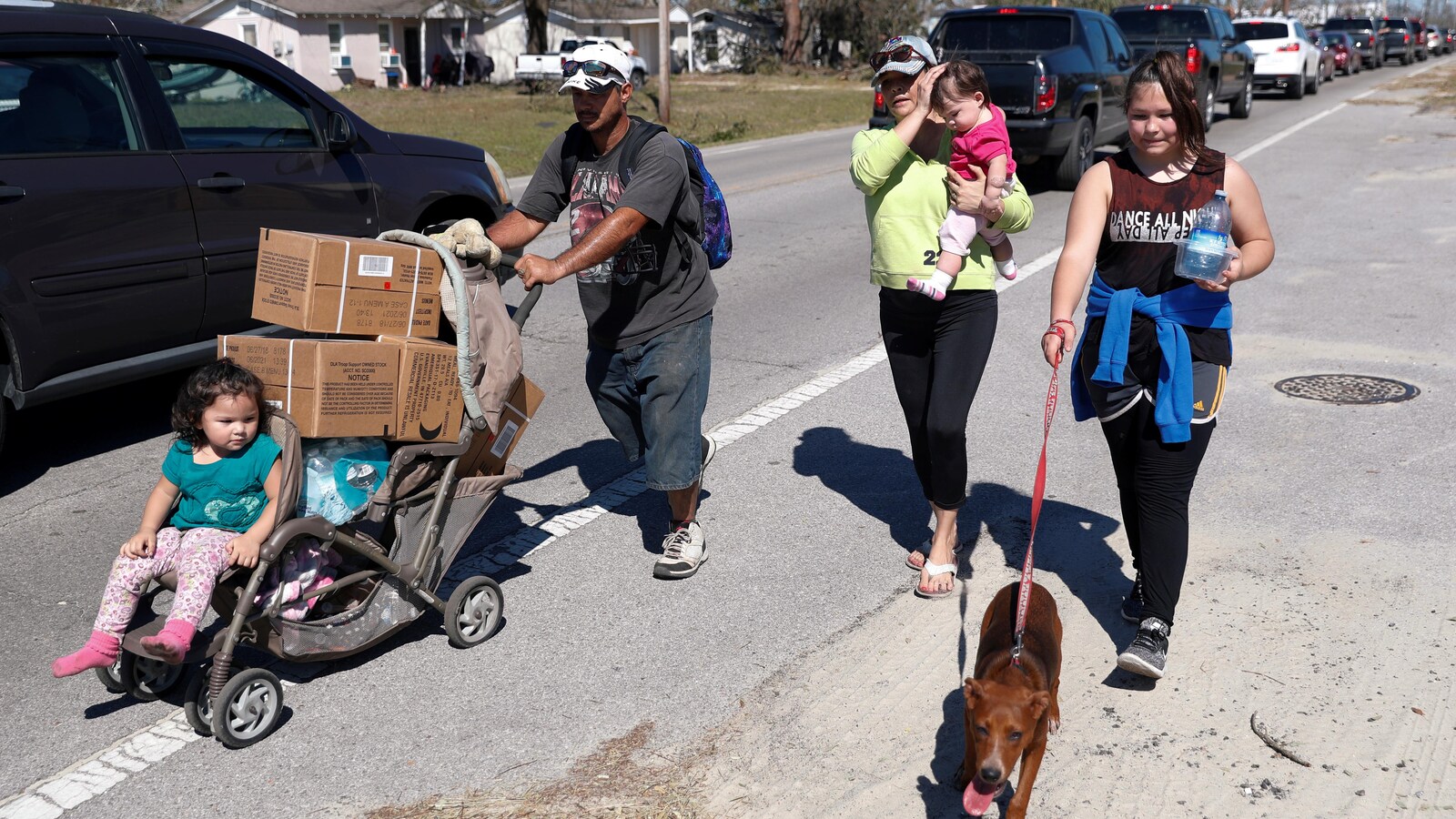 Un homme pousse une poussette avec un enfant et plusieurs boîtes de vivres. Une femme avec un bébé dans les bras et une adolescente avec un chien en laisse l'accompagnent. Une longue fil de voiture est immobilisée sur la route derrière eux.