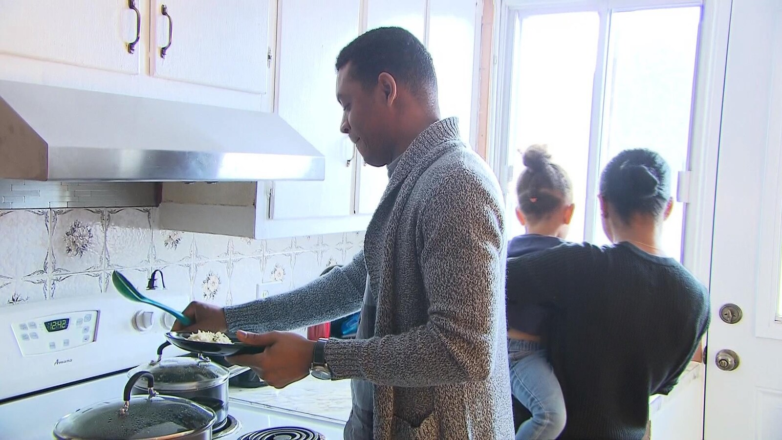 Endy Labaty cuisine, pendant que sa femme Andie regarde dehors en tenant leur fillette dans ses bras.
