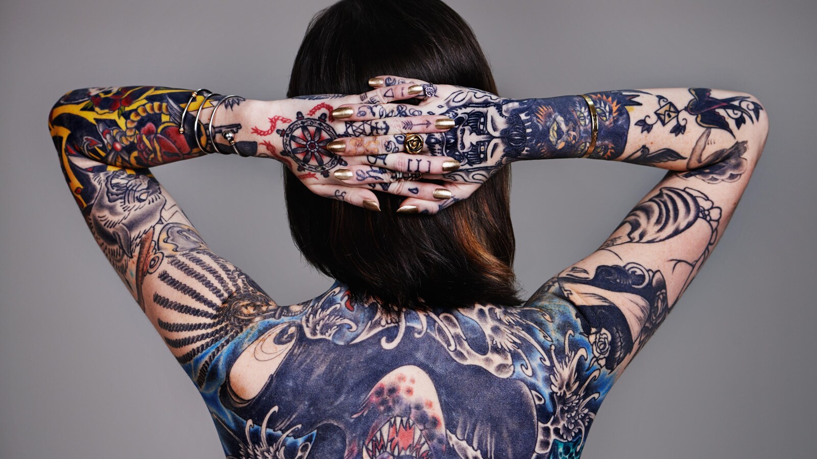 On voit une femme, de dos, les mains jointes derrière la tête, qui expose les tatouages qu'elle a sur son corps.
