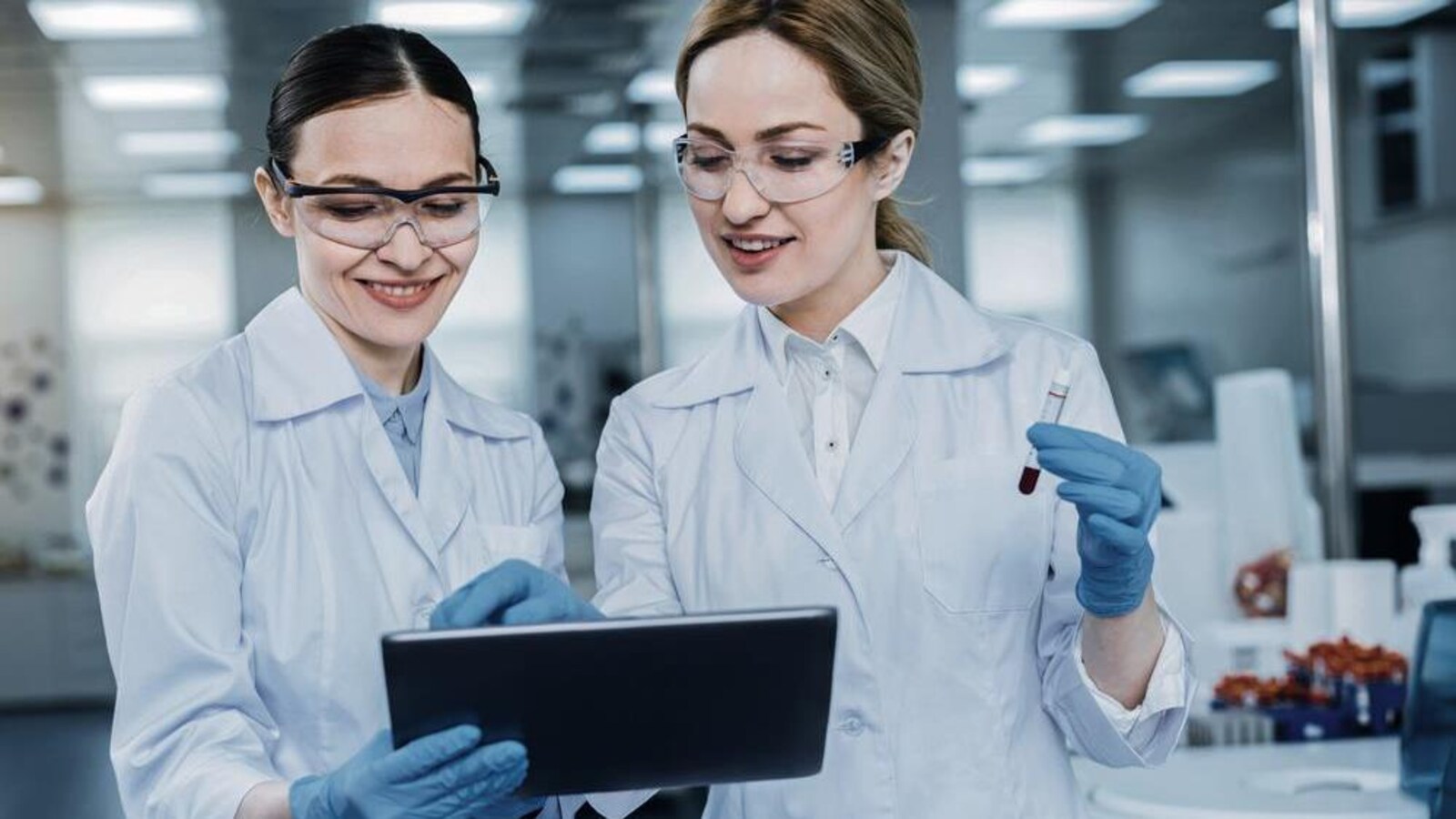 Deux femmes regardent une tablette électronique. Elles portent des gants, des sarraus et des lunettes. Une tient une éprouvette.
