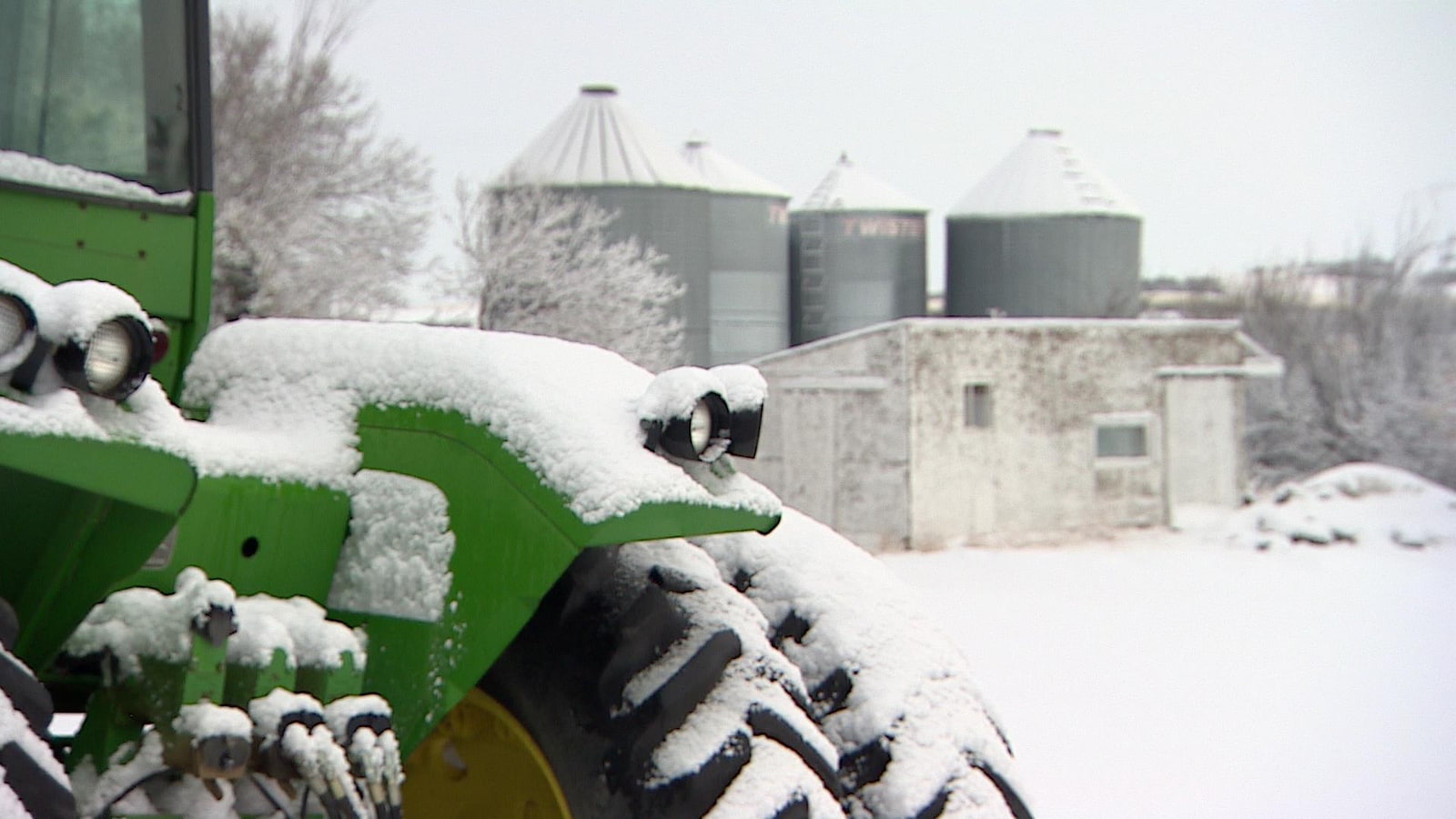 Un tracteur vert enneigé devant un cabanon et des installations agricoles, en temps de neige.