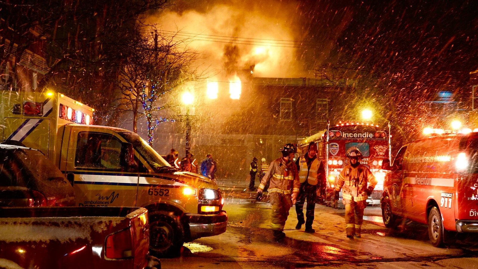 Vue d'ensemble de la scène : ambulance, camion de pompiers, pompiers, flammes qui sortent par les fenêtres de l'immeuble.