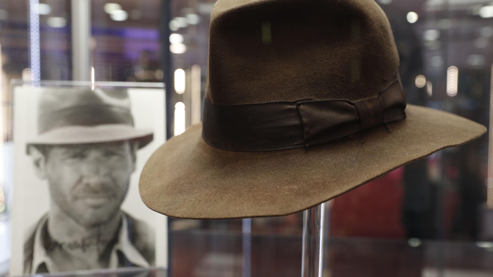 On voit le chapeau sur un support avec la photo de l'acteur qui le porte en arrière-plan.