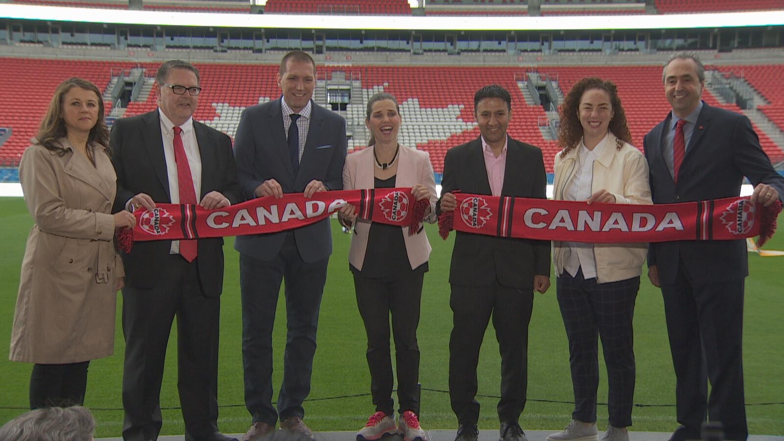 la ministre fédérale des sports Kirsty Duncan est avec des membres de l'Association canadienne du soccer et une banderole avec écrit dessus « Canada » devant le BMO Field de Toronto, ils sourient et posent pour une photo de groupe.