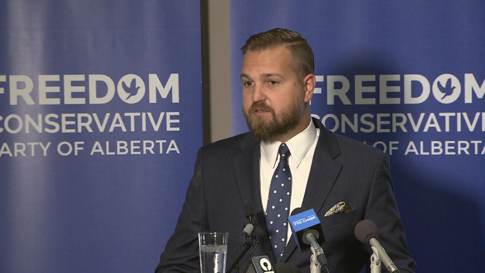 Un homme barbu en veston cravate est debout devant deux affiches où il est écrit en anglais : «Parti conservateur libre de l'Alberta ». Devant lui, plusieurs micros.