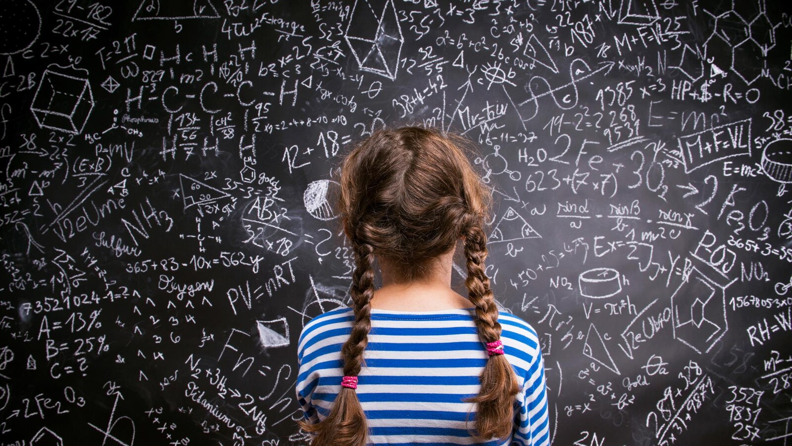 Une fille de dos qui regarde un grand tableau noir avec des formules mathématiques.
