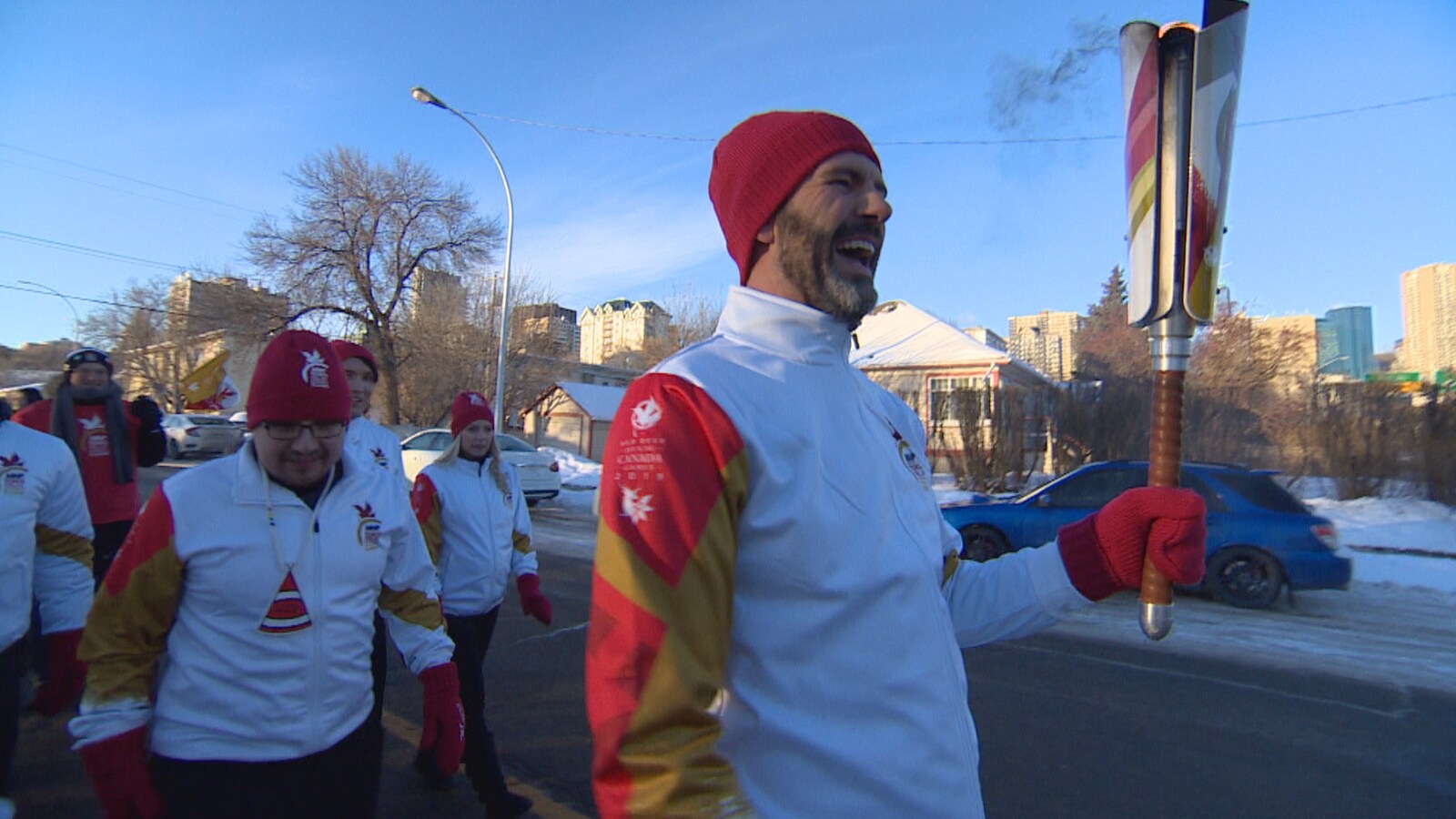 Don Iveson porte un flambeau, dehors. Il semble être en train de rire. Une petite foule habillée aux couleurs des Jeux du Canada le suit.