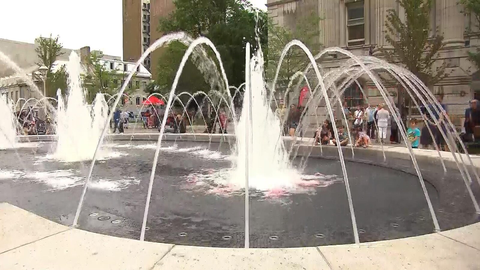 La place Vauquelin rénovée, à côté de l'hôtel de ville, à Montréal, dont on voit la fontaine, entourée de gens.