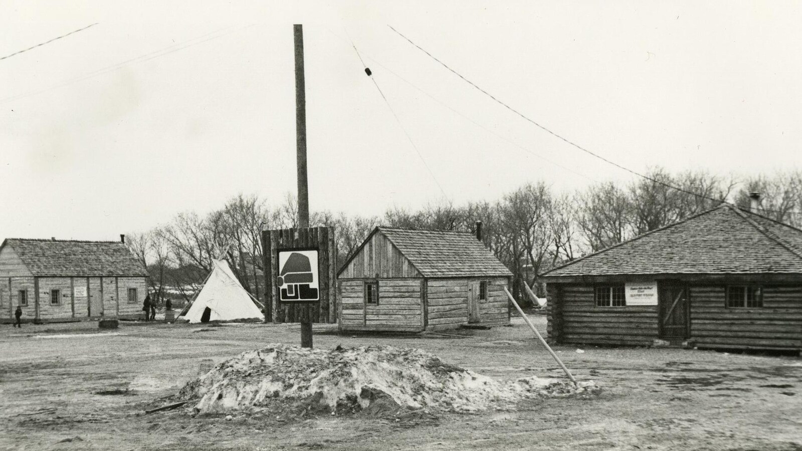 4 cabanes de bois rond dans la neige. Entre la deuxième et la troisième se dresse un tipi.