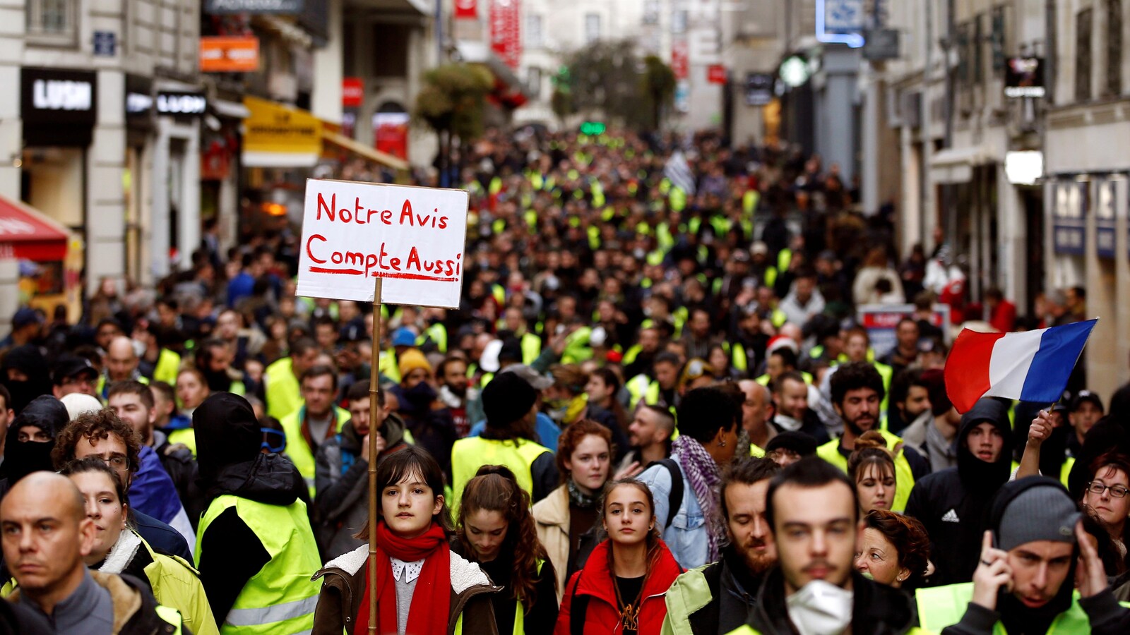 Une marée de gens occupe une rue. Tout le monde porte un gilet jaune. Un femme tient une pancarte sur laquelle est écrit : « Notre avis compte aussi ».
