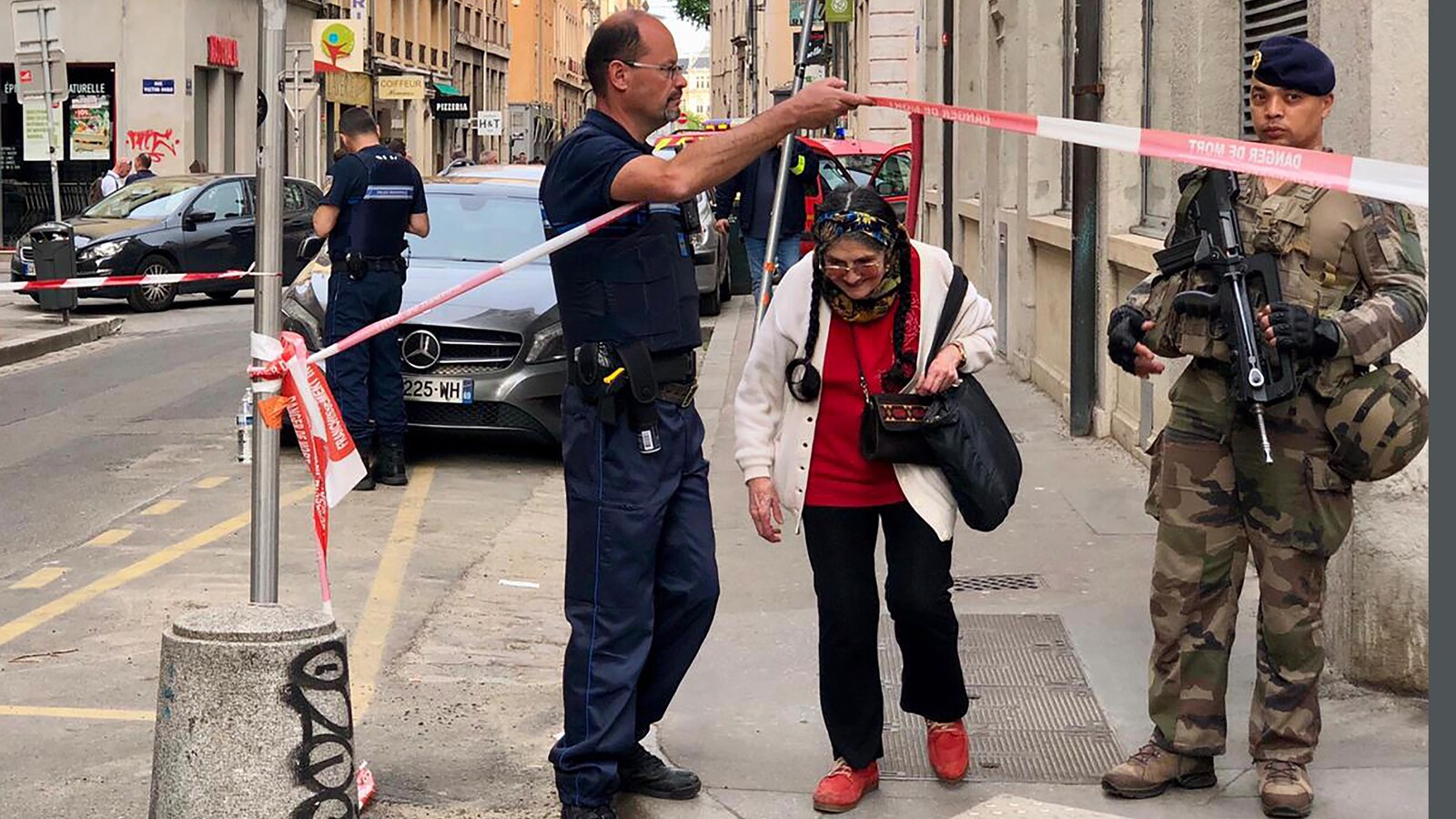 Dans une rue commerciale, un policier laisse passer une vieille dame sous un cordon de sécurité.