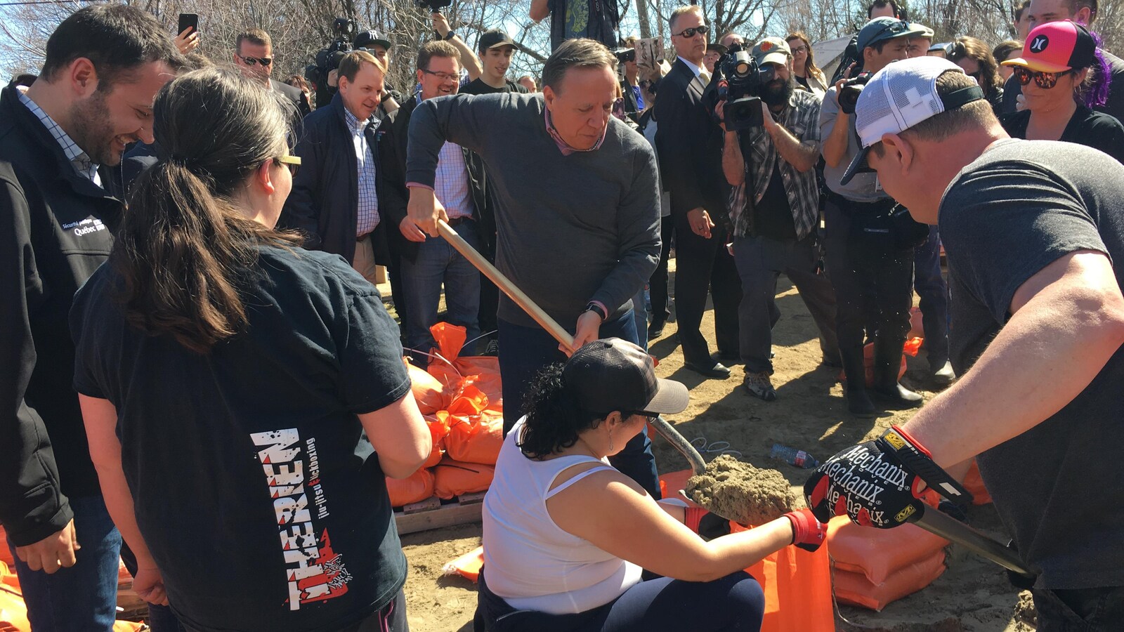 M. Legault pellette du sable dans un sac, entouré de bénévoles, de politiciens et de représentants des médias.