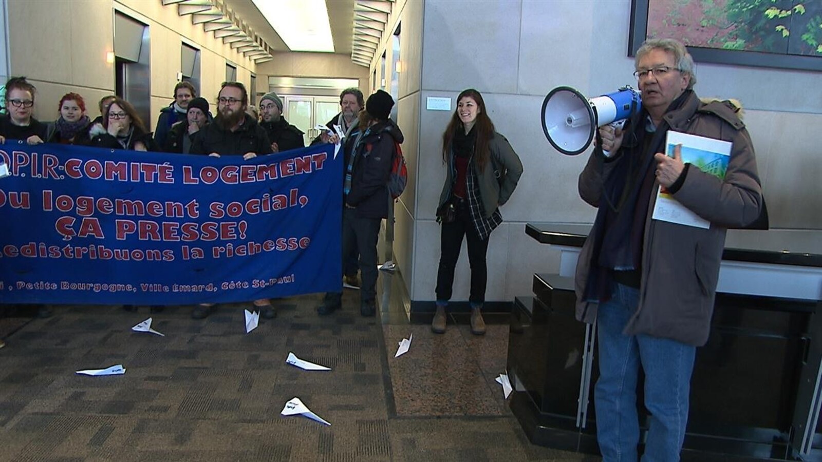 Un homme portant un manteau parle dans un mégaphone devant des manifestants tenant une banderole dans le hall à l'entrée d'un immeuble.