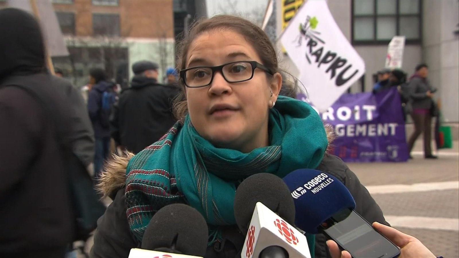 Émilie Joly est photographiée devant les micros de différents médias; derrière elle, des manifestants tiennent des pancartes réclamant des logements sociaux.