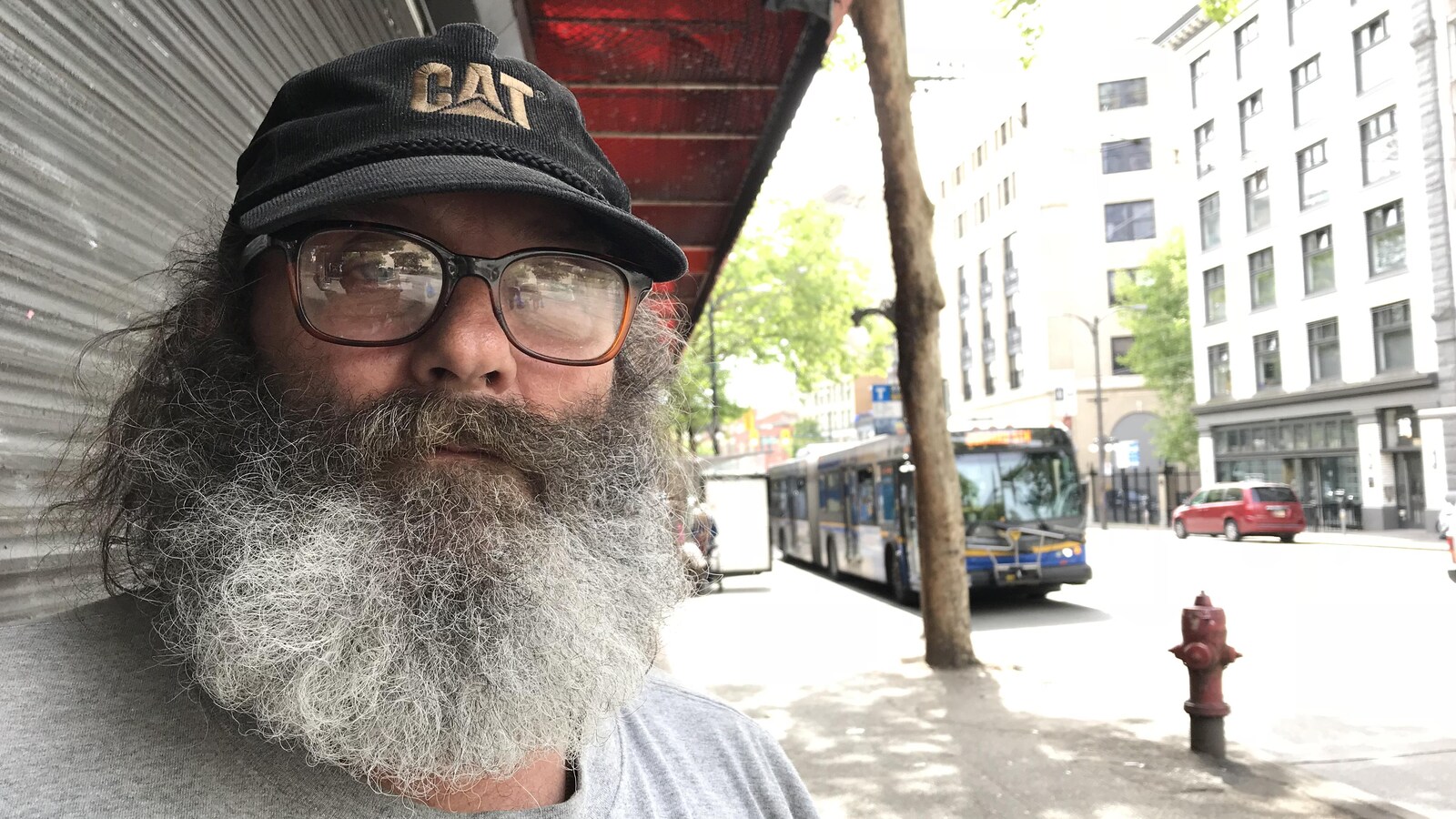 Un homme à la barbe foisonnante poivre et sel arbore une casquette noire et des lunettes à épaisses montures. Il se trouve sur un trottoir du centre-ville.