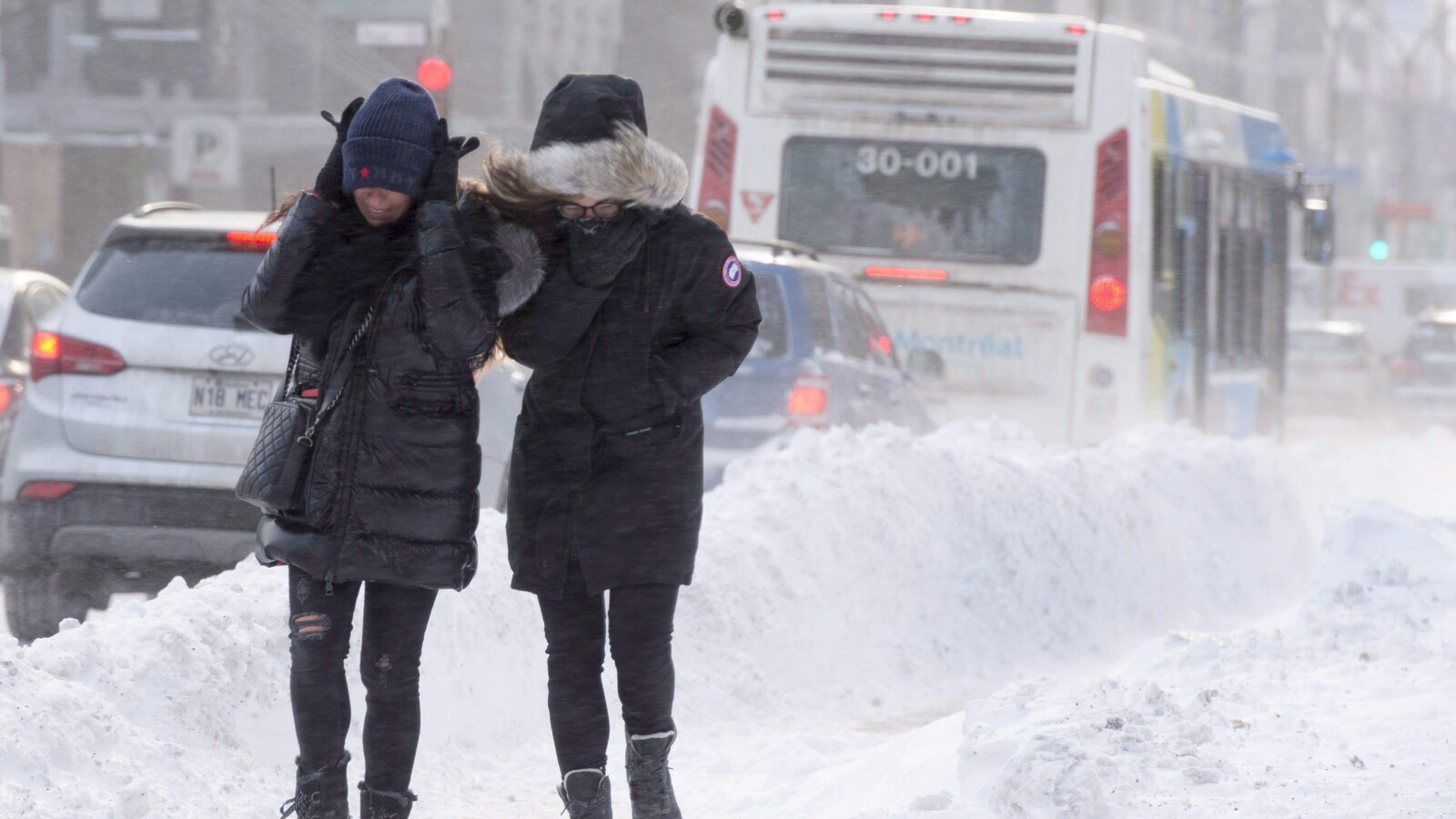 Deux personnes marchent sur un trottoir enneigé et se protègent du vent glacial. Elles sont lourdement habillées.