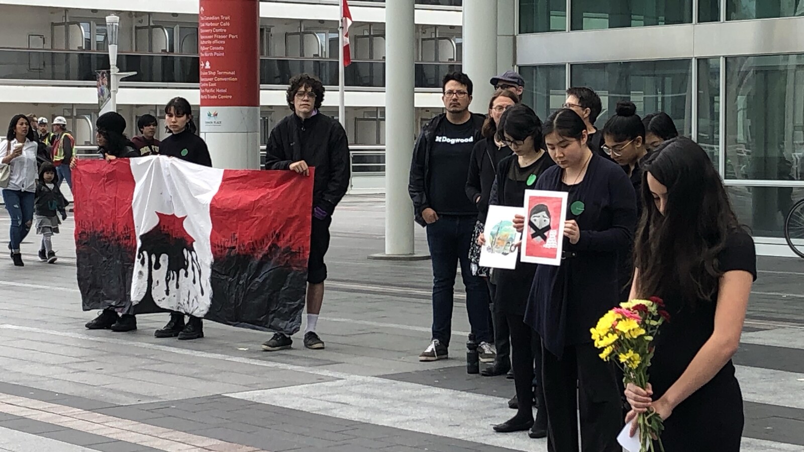 Des adolescents habillés en noir, certains la tête baissée, avec des fleurs ou des affiches dans les mains, à gauche trois jeunes qui tiennent un drapeau du Canada avec la feuille à l'envers, qui dégouline d'huile.