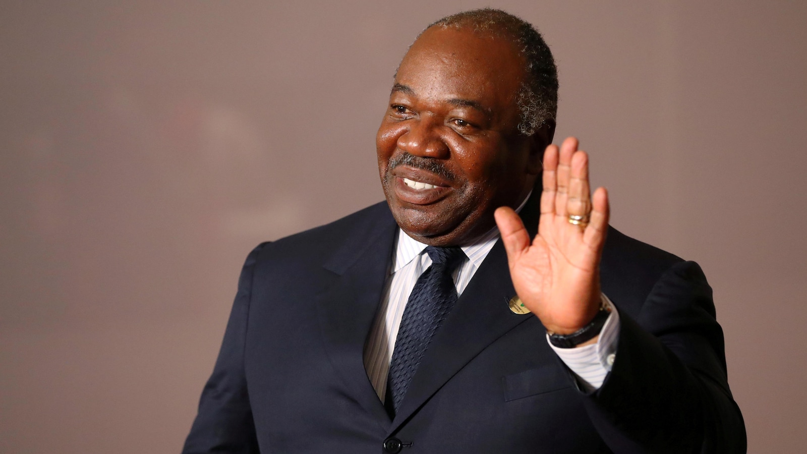Le président gabonais, portant un complet et une cravate, salue de la main en souriant.