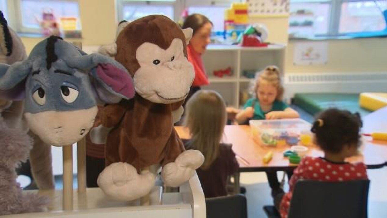Des jouets sont à l'avant-plan de la photo qui montrent aussi une éducatrice assise à une table avec des enfants