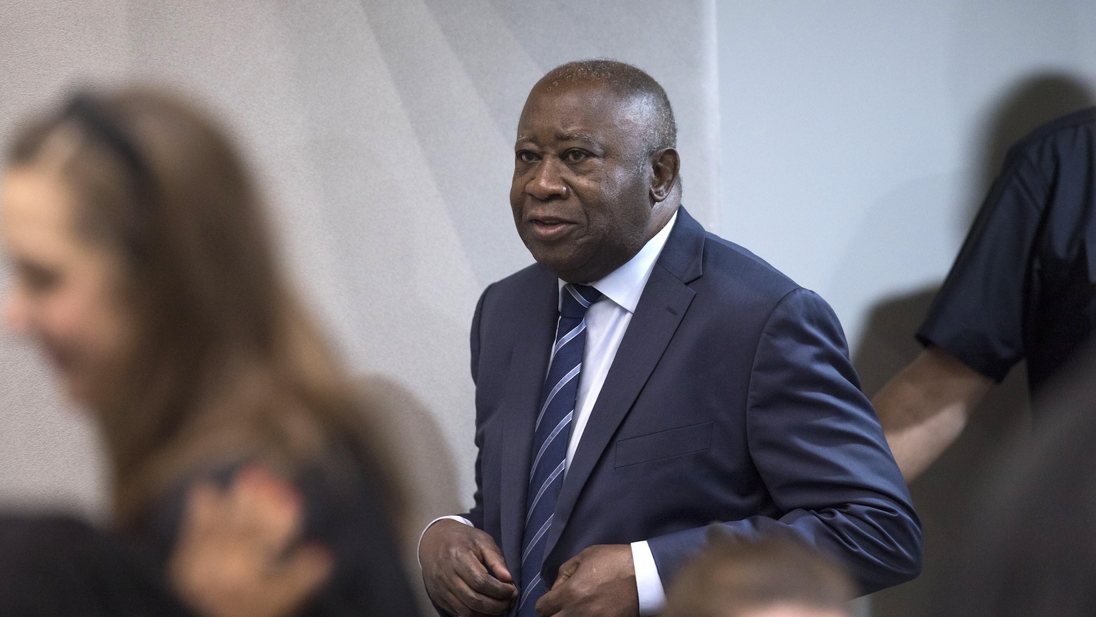 Un homme ajuste son veston, entouré de plusieurs personnes dans un corridor aux murs blancs.