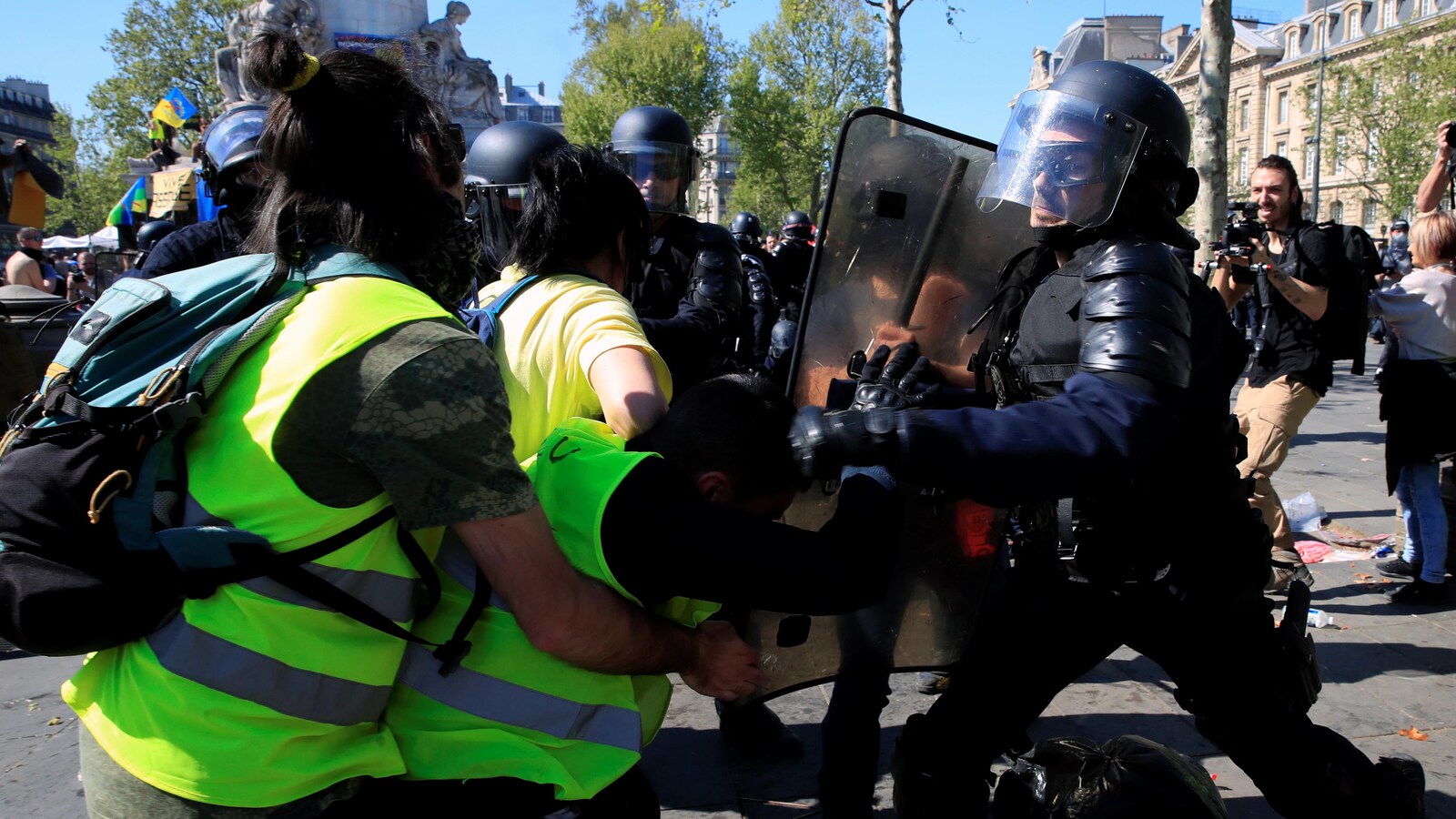 Des gilets jaunes se font repousser par un policier casqué, qui porte un bouclier.