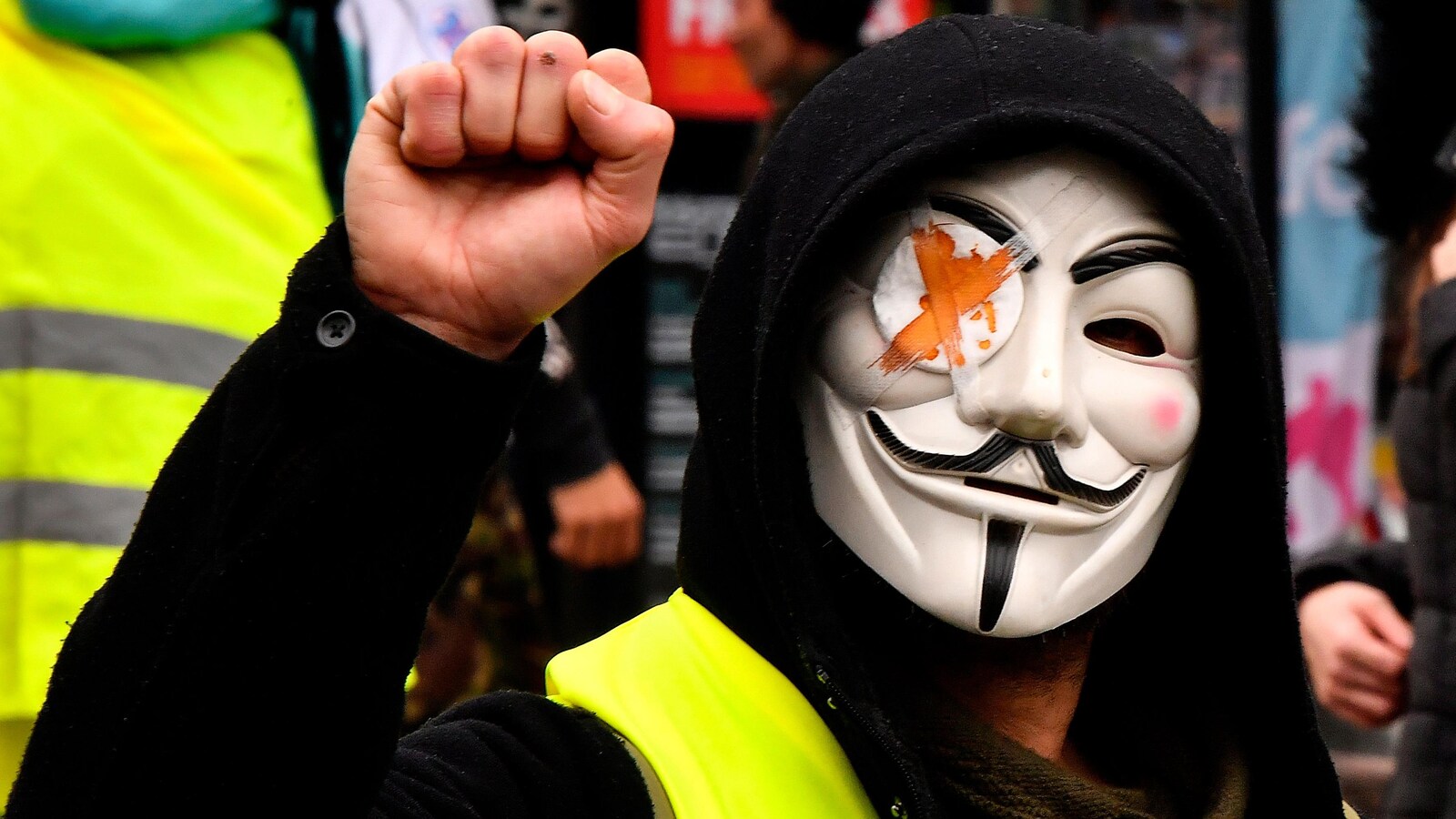 Un manifestant portant un gilet jaune, un masque d'Anonymous avec un bandeau sur l’œil droit brandit le poing.