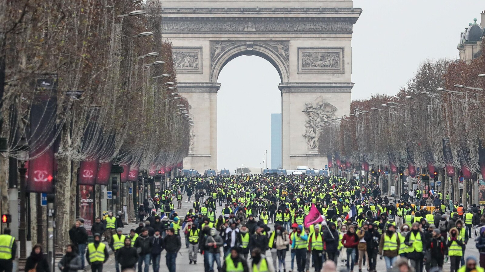 Plusieurs centaines de personnes marchent à Paris, plusieurs portant un gilet jaune. À l'arrière se dresse l'Arc de Triomphe.