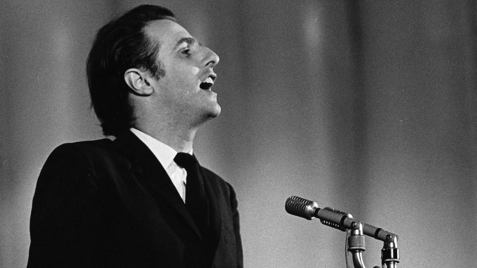 Sur une scène, Gilles Vigneault, chante, un micro sur pied devant lui.