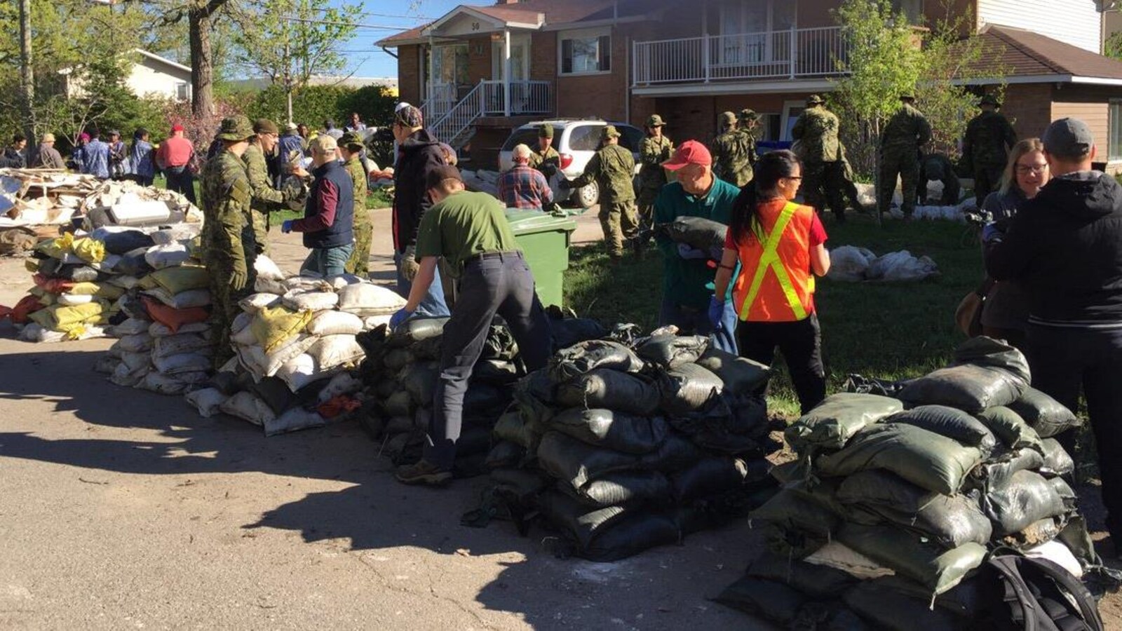 Des militaires et des bénévoles ramassent des sacs de sable devant une maison.