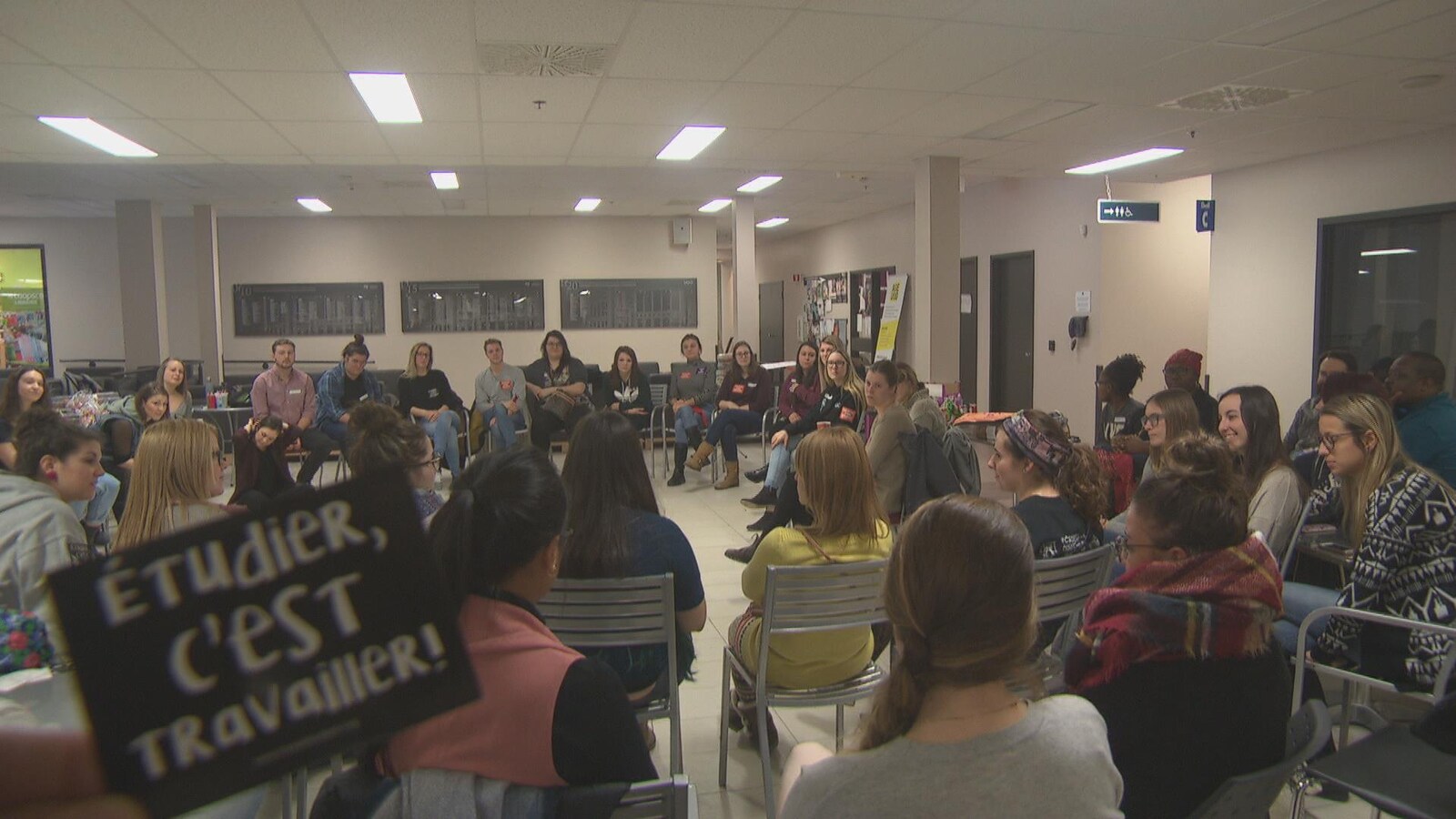 Les étudiants sont assis en cercle et écoute des intervenants. Le caméraman tient dans un coin un carton sur lequel est écrit : étudier, c'est travailler.