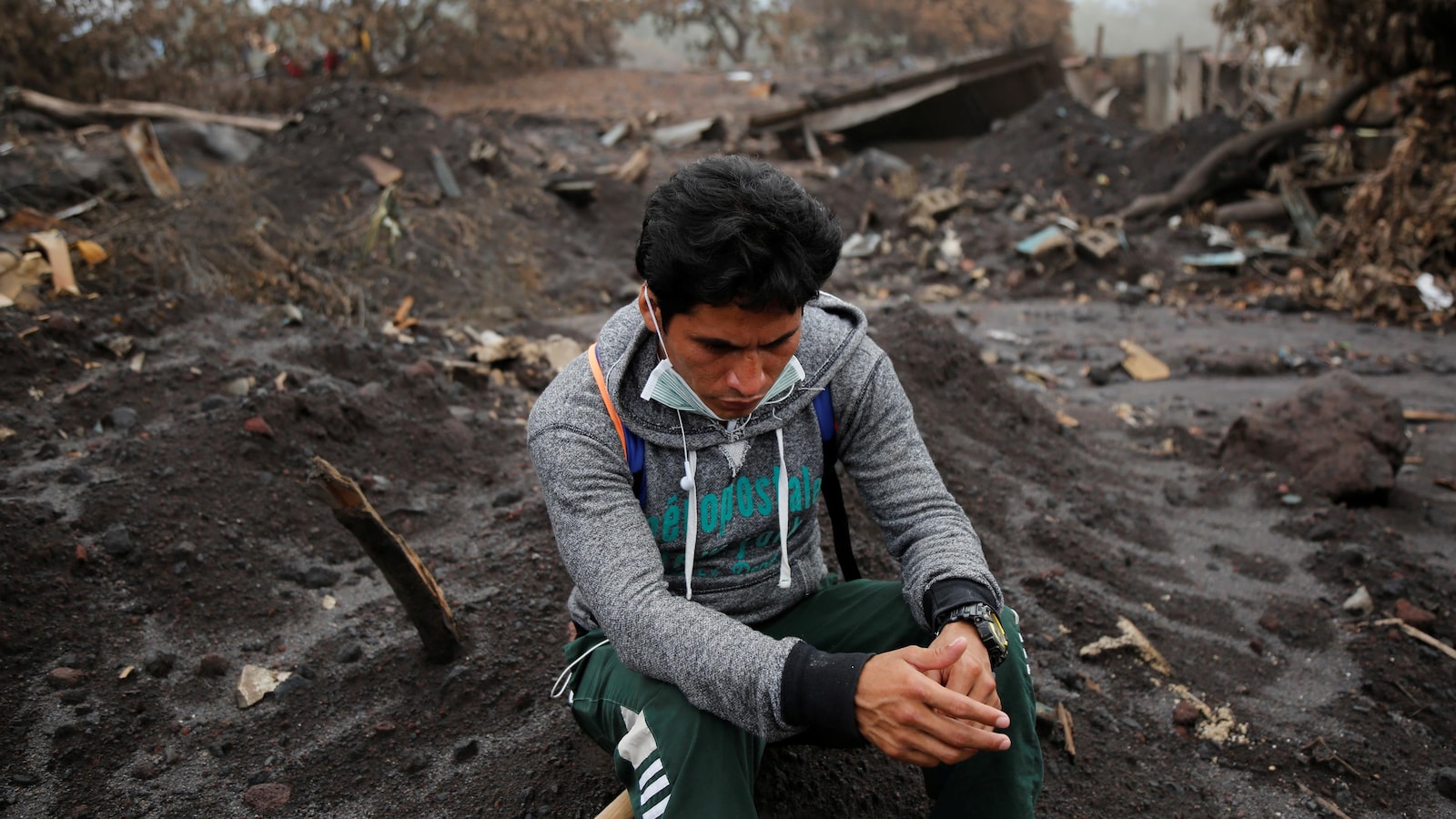 Un homme qui fouille les décombres après l'éruption du volcan de Fuego prend une pause. Le sol est ravagé et les bâtisses en arrière-plan sont démolies.