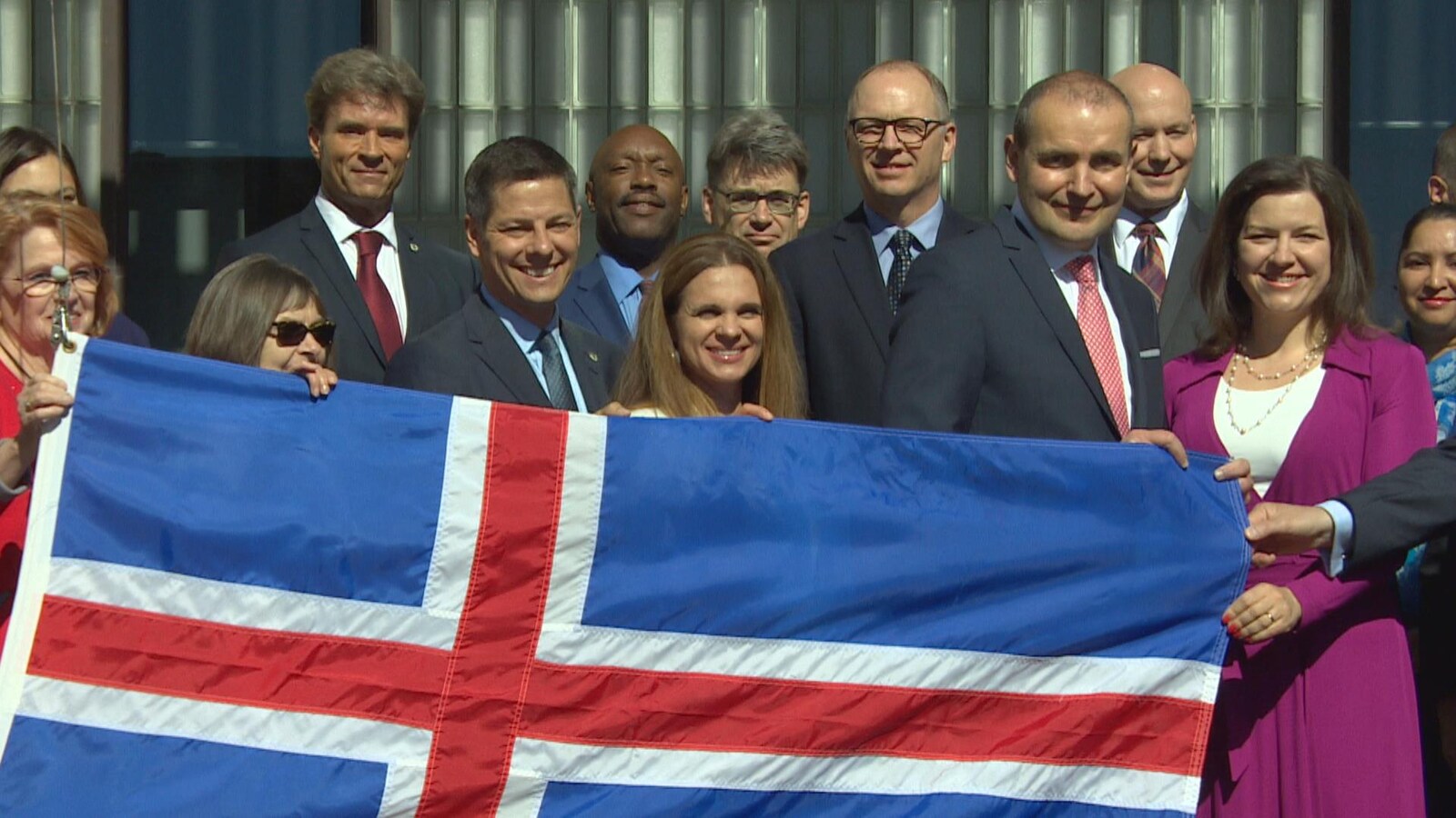 Une dizaine de personnes en complets et vêtements chics, derrière un grand drapeau islandais. Les personnes à l'avant-plan (le maire, le président islandais et leurs épouses respectives) tiennent le haut du drapeau.
