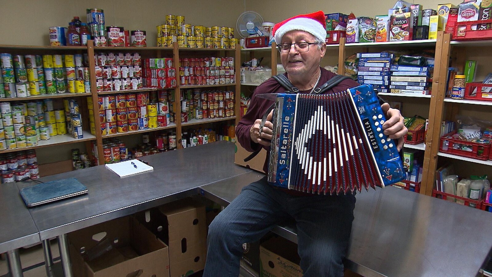 Un bénévole de La guignolée joue de l'accordéon avec une tuque du Père Noël devant des étagères pleines de denrées