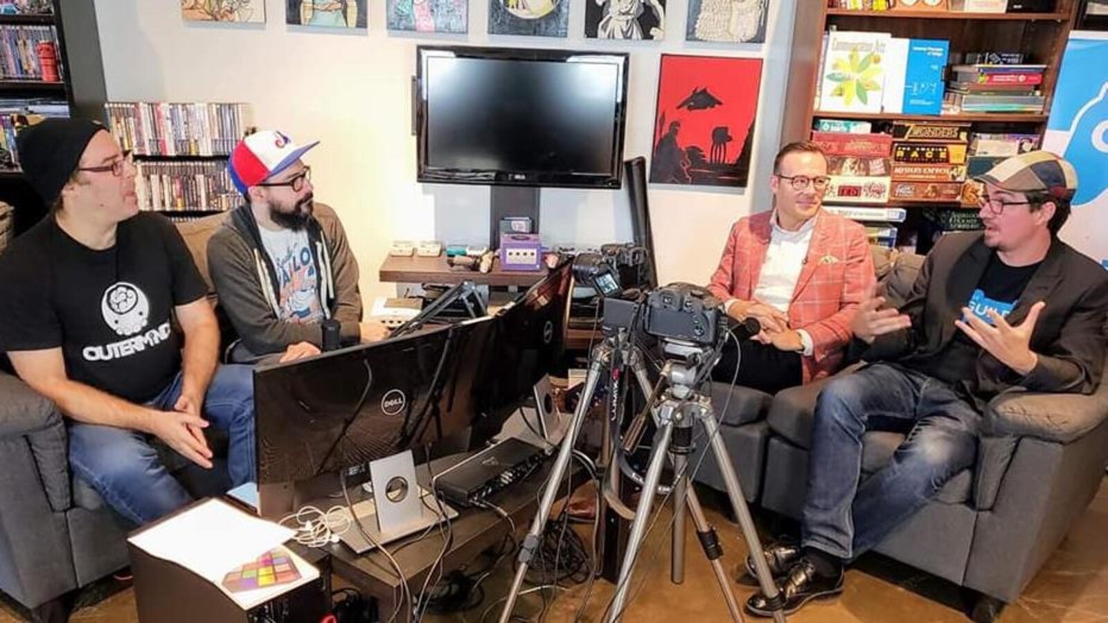 Quatre hommes discutant sur des sofas entourés. Des appareils électroniques, des jeux de société et des jeux vidéo occupent une bonne partie de l'espace et des étagères les entourant.