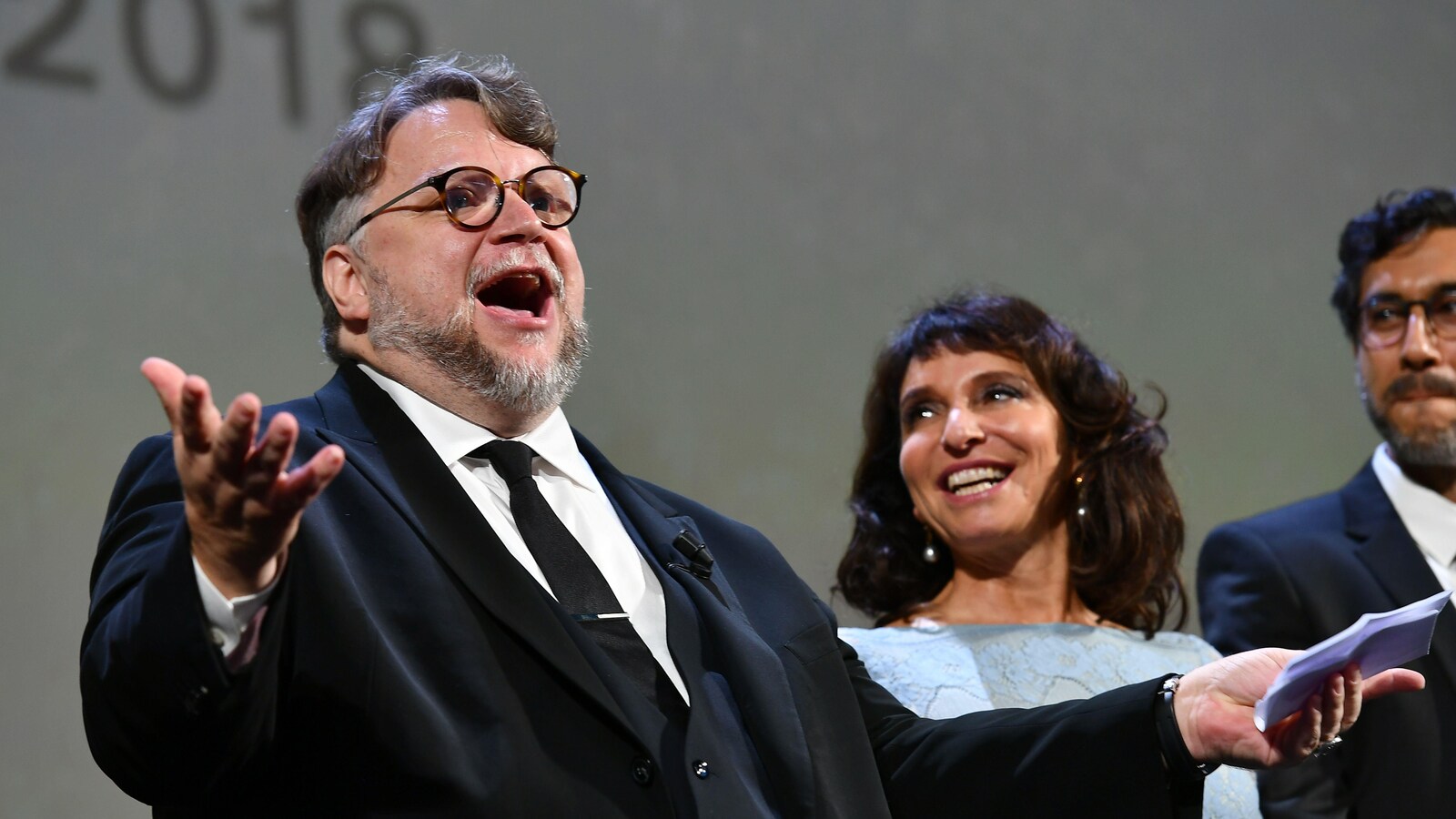 L'homme porte des lunettes et une barbe, il a les bras allongés devant lui pendant que les deux autres personnes le regardent.