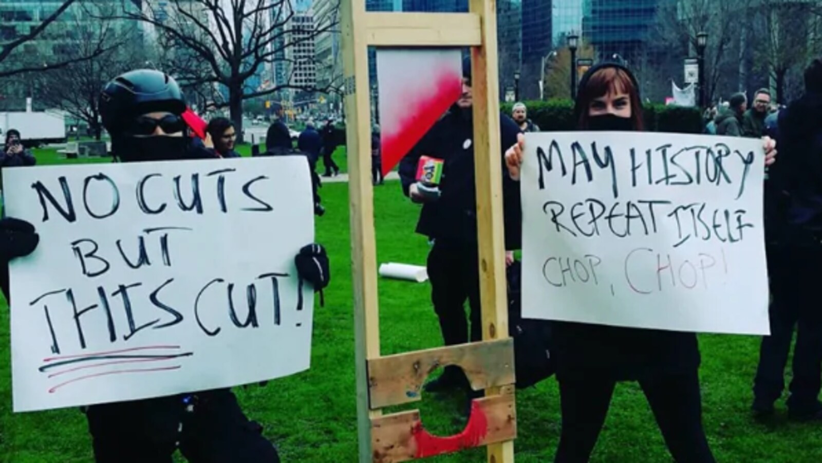 Des manifestants vêtus de noir et masqués tiennent des cartons où on peut lire des messages dénonçant les compressions.