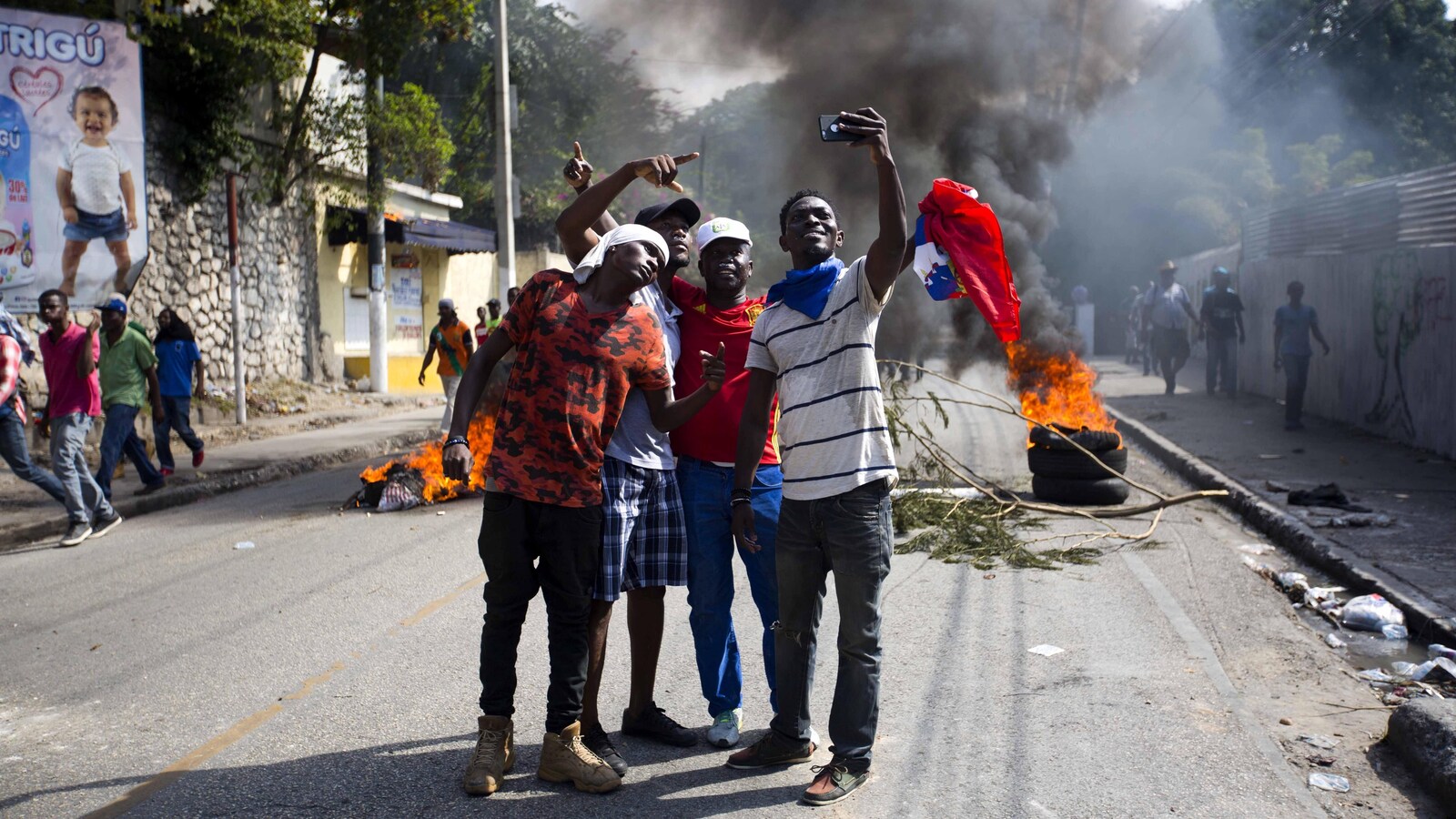 Quatre jeunes Haïtiens se prennent en photo avec un téléphone portable dans une rue où se déroule une manifestation avec des pneus en feu et une épaisse fumée.