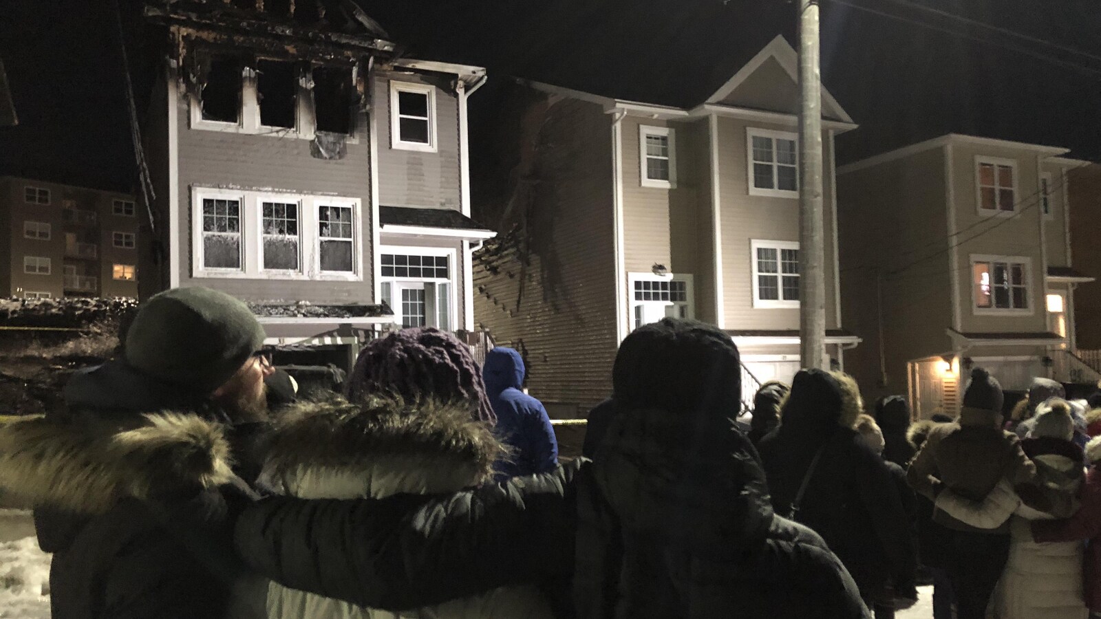 Les gens rassemblés devant la maison ravagée par les flammes se tiennent bras dessus bras dessous.