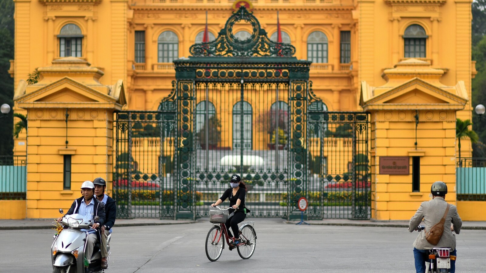 Deux cyclistes et deux personnes sur une moto circulent devant le palais présidentielle à Hanoï.