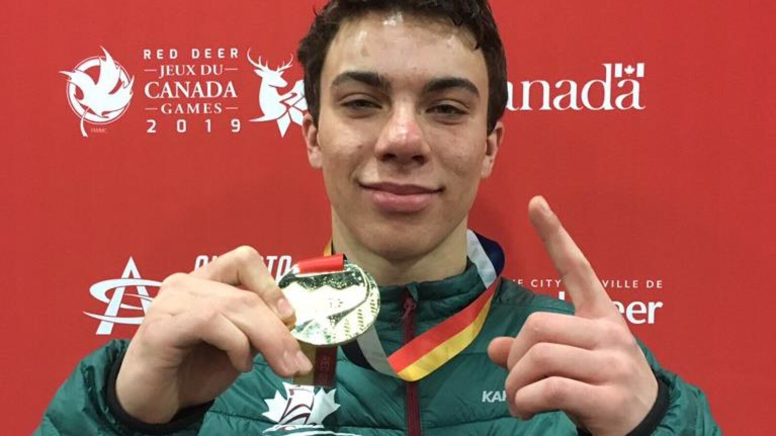 L'athlète, souriant, tient sa médaille d'une main, et il fait un signe de victoire de l'autre main.