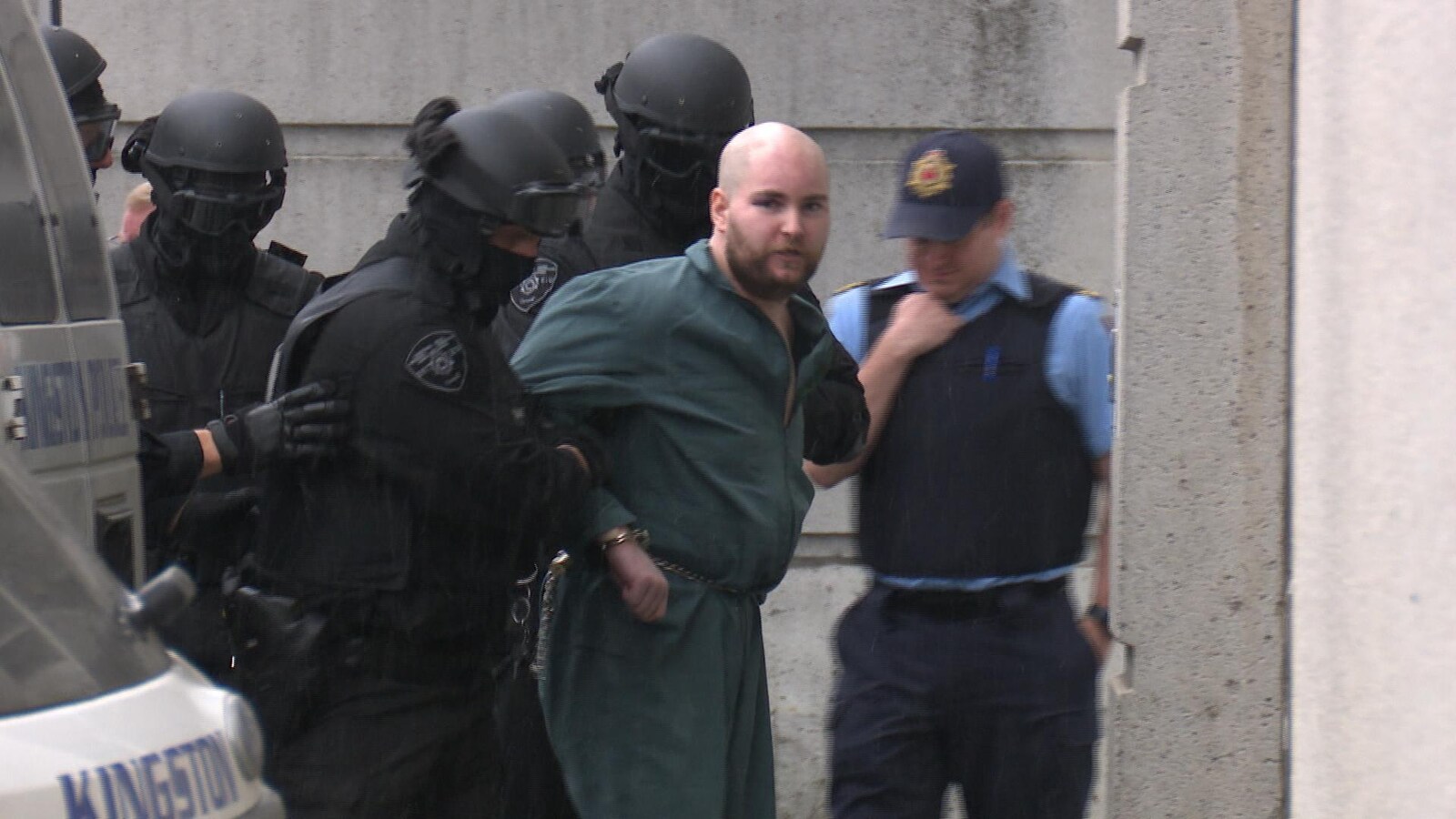 Photo d'un homme menotté habillé en vert qui est escorté par des policiers vêtus de noir.